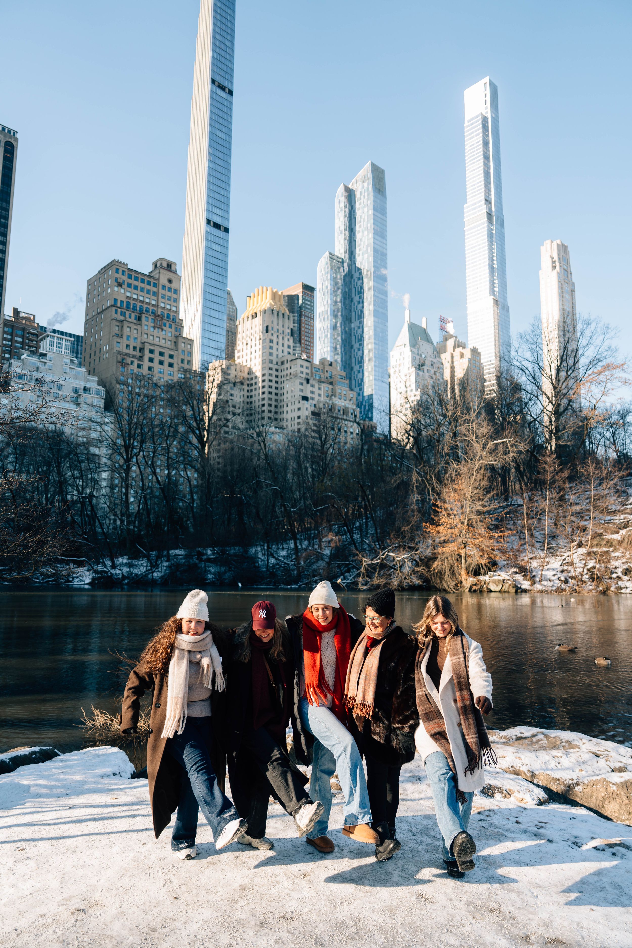 Five friends walk along a snowy Central Park path with Manhattan skyscrapers reflecting in a pond on a sunny winter day.