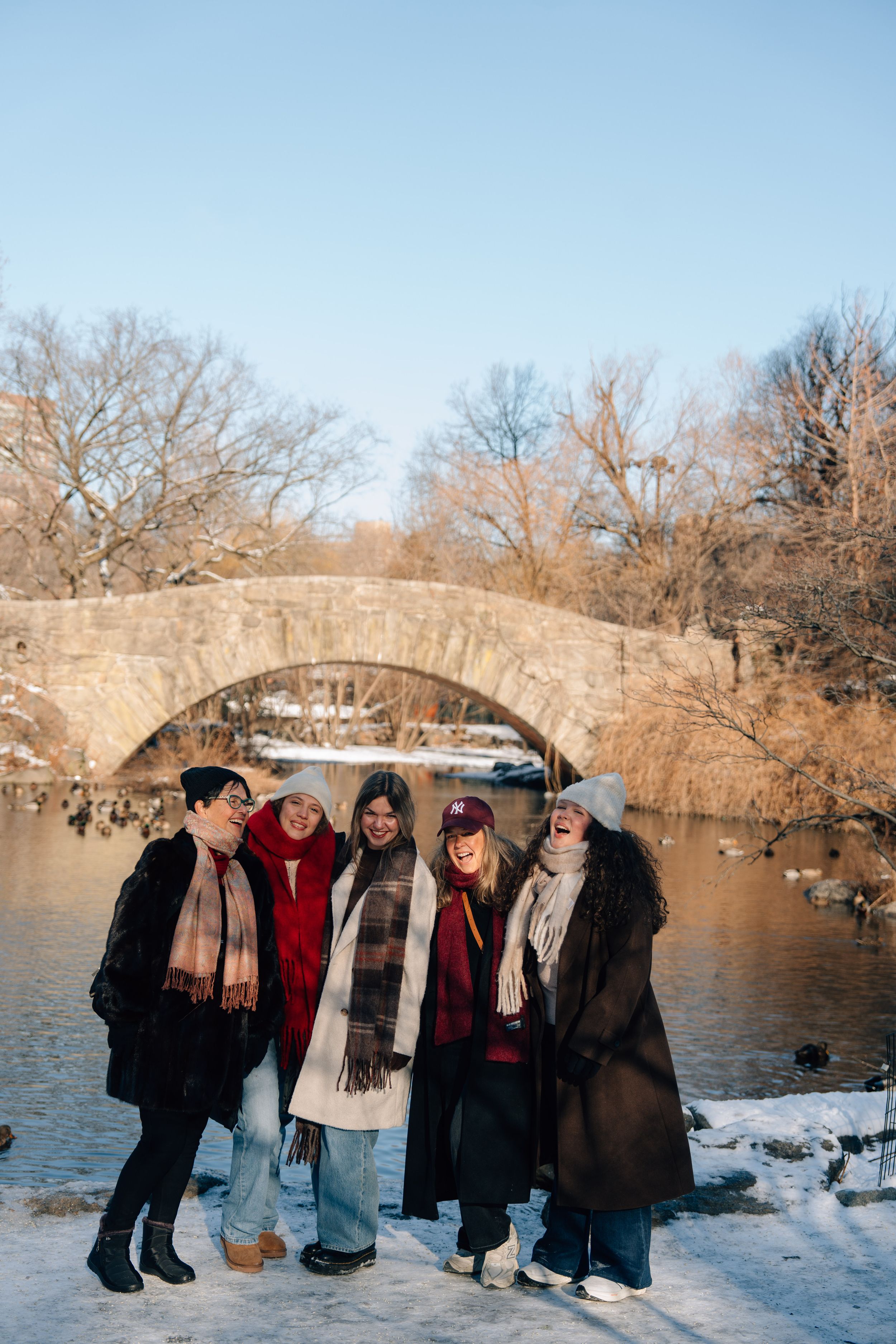 Five women in winter coats and scarves laugh together by a snowy Central Park pond with a stone bridge backdrop.