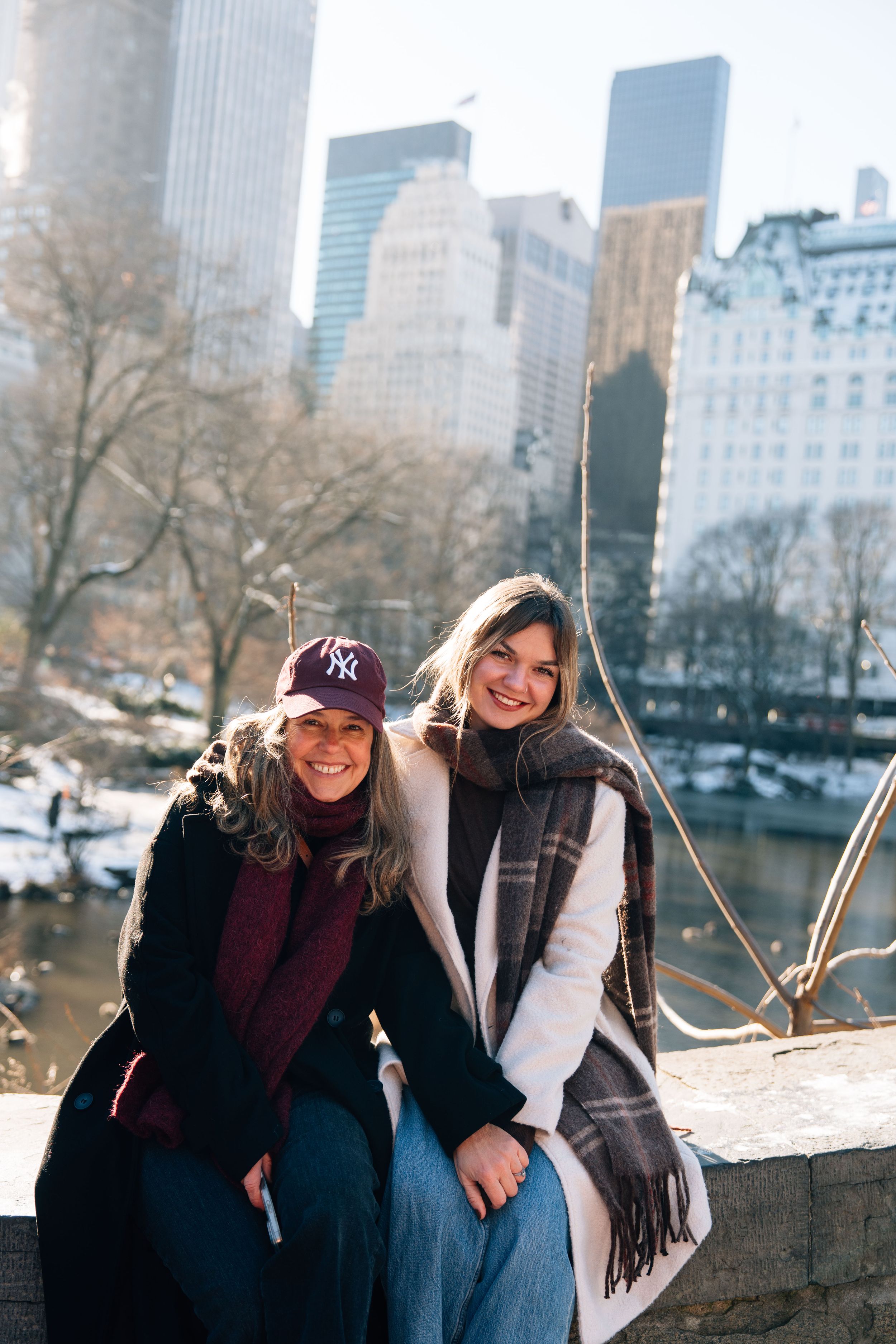 Two women smiling together on a bridge in Central Park, NYC, with city skyline and pond in winter background.