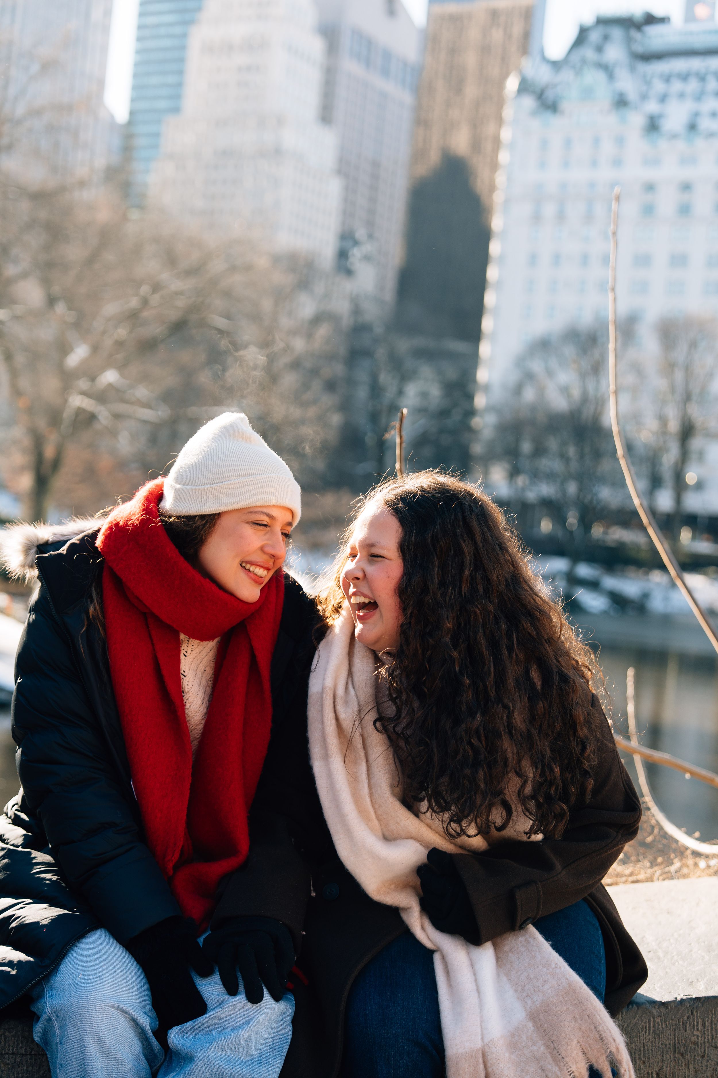 Two women laughing together in winter scarves and coats with NYC skyline and Central Park in background.