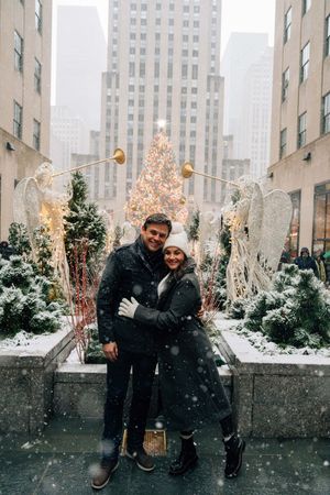 Couple embracing in front of Rockefeller Center Christmas tree during snowfall in New York City.