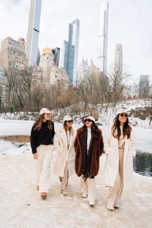 Four women in winter coats walk through snowy Central Park with NYC skyscrapers in the background.
