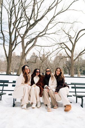 Four women in winter coats sit on a snowy Central Park bench, smiling and wearing sunglasses on a cold day.
