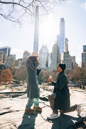 Man proposes on one knee to woman in Central Park with NYC skyline in background on a sunny winter day.