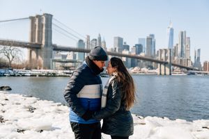 Couple sharing a romantic moment by the Brooklyn Bridge with NYC skyline and icy waterfront in winter.
