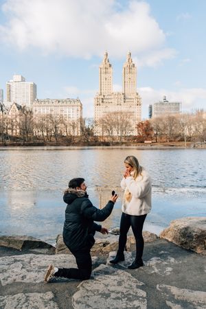 Man proposes to woman on one knee by a lake in Central Park, NYC, with iconic skyline buildings in the background.