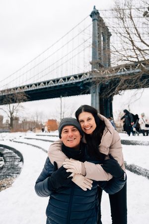 Couple embracing in the snow at Brooklyn Bridge Park, NYC, wearing winter coats and hats on a cloudy day.