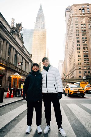 Two young men posing on a crosswalk in New York City with the Chrysler Building visible in the background.