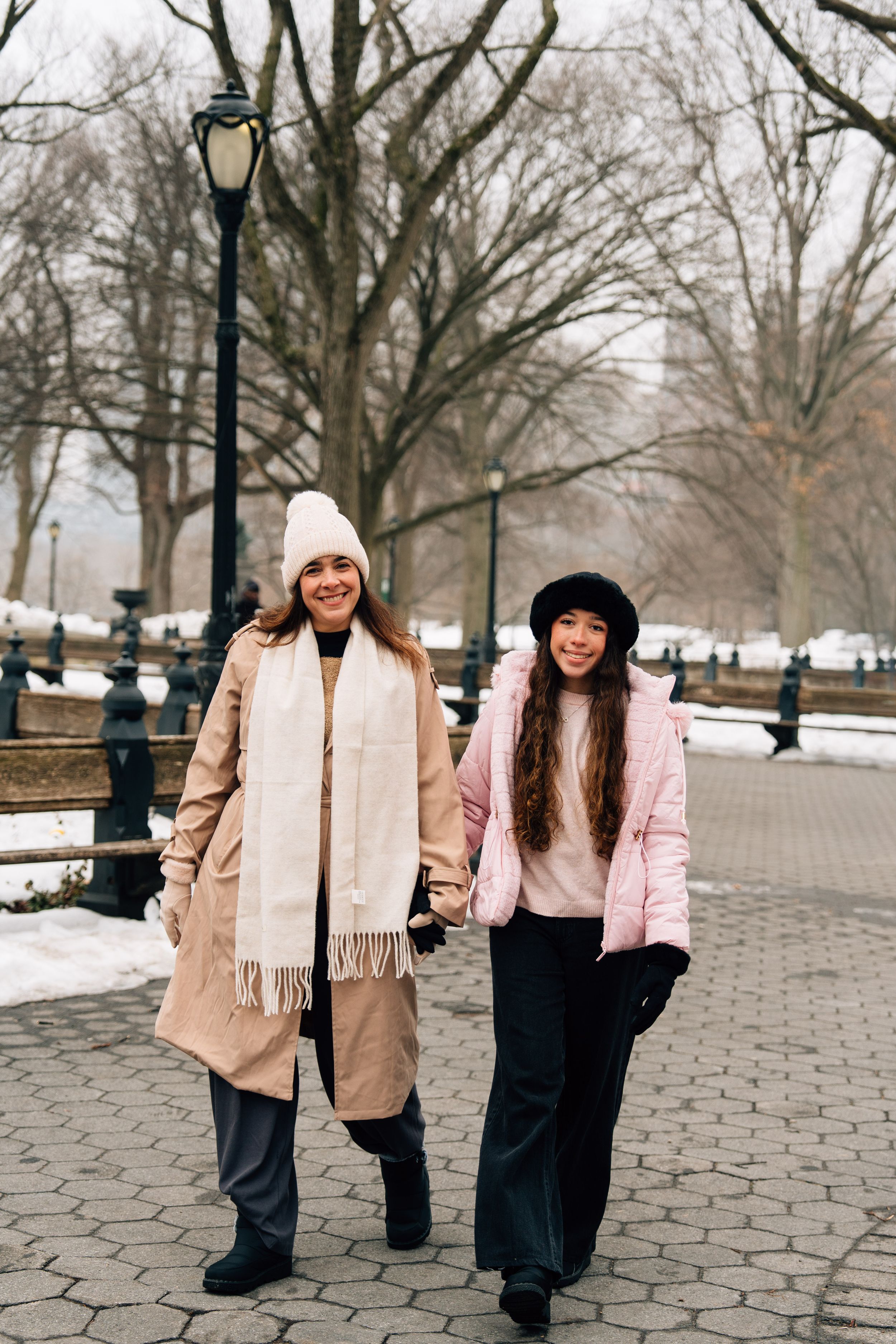 Two women in winter coats and hats walk hand in hand through a snowy Central Park in New York City.