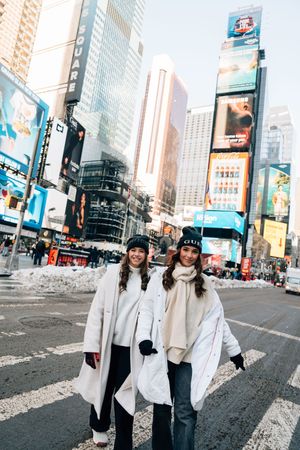 Two women in white winter coats and black beanies smiling in Times Square, New York City on a snowy day.