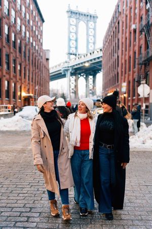 Three women walking on cobblestone street in DUMBO Brooklyn with Manhattan Bridge in background on snowy winter day.
