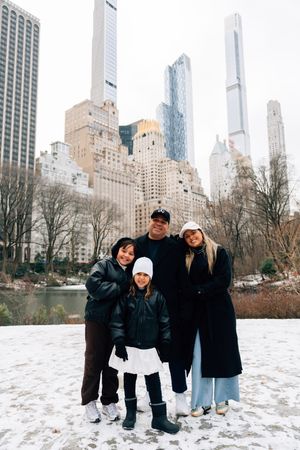 Family of four posing in snowy Central Park with NYC skyline skyscrapers in the background on a winter day.