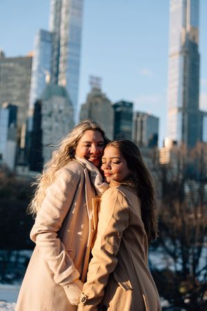 Two women in beige coats smile together in Central Park with NYC skyline skyscrapers in the snowy winter background.