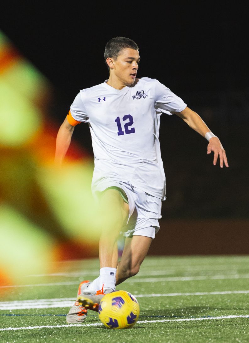 High school soccer player wearing number 12 dribbling the ball during a nighttime game.