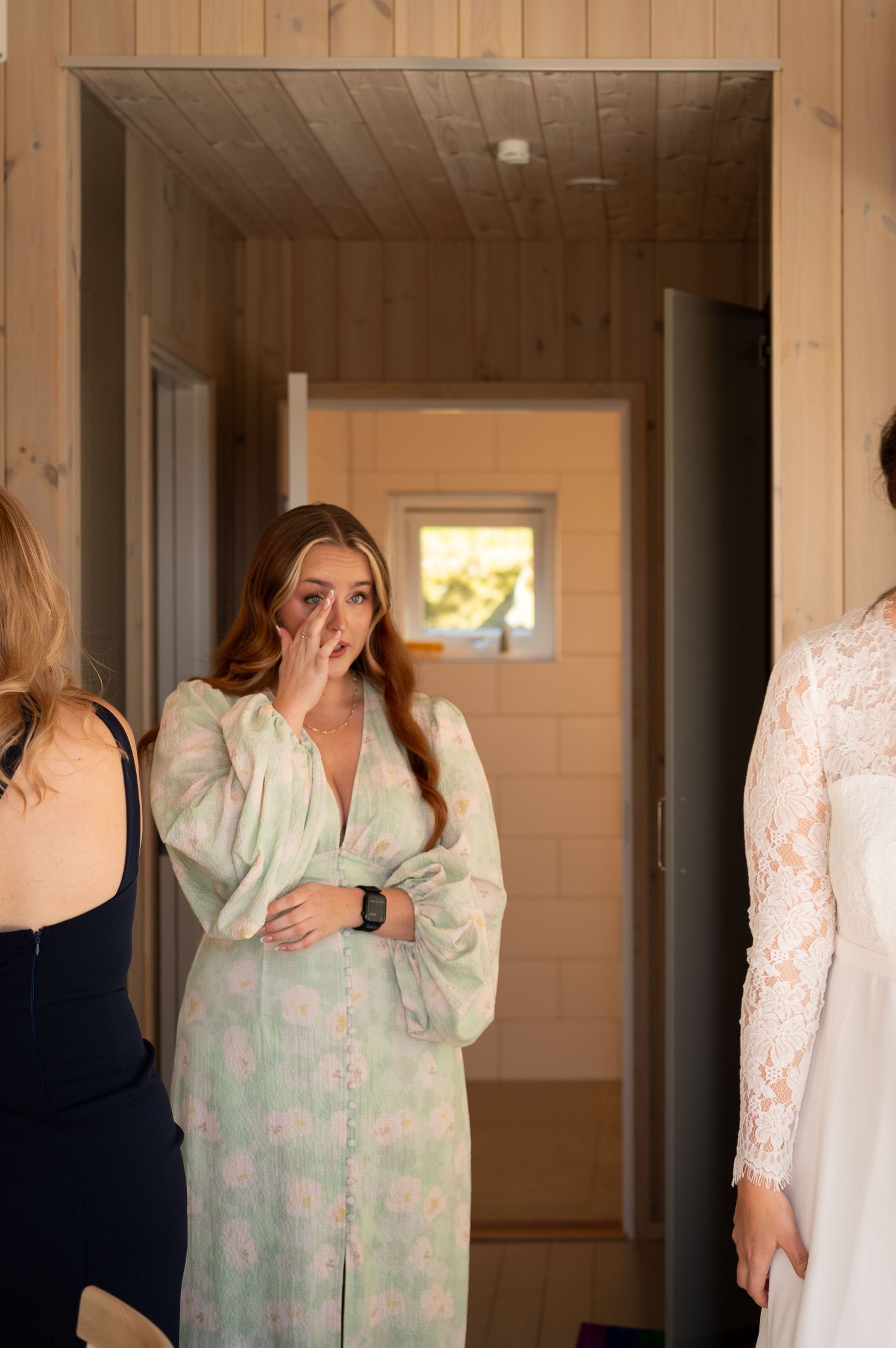 A bridesmaid in a floral robe wipes away tears while viewing the bride in her wedding dress during bridal preparations.
