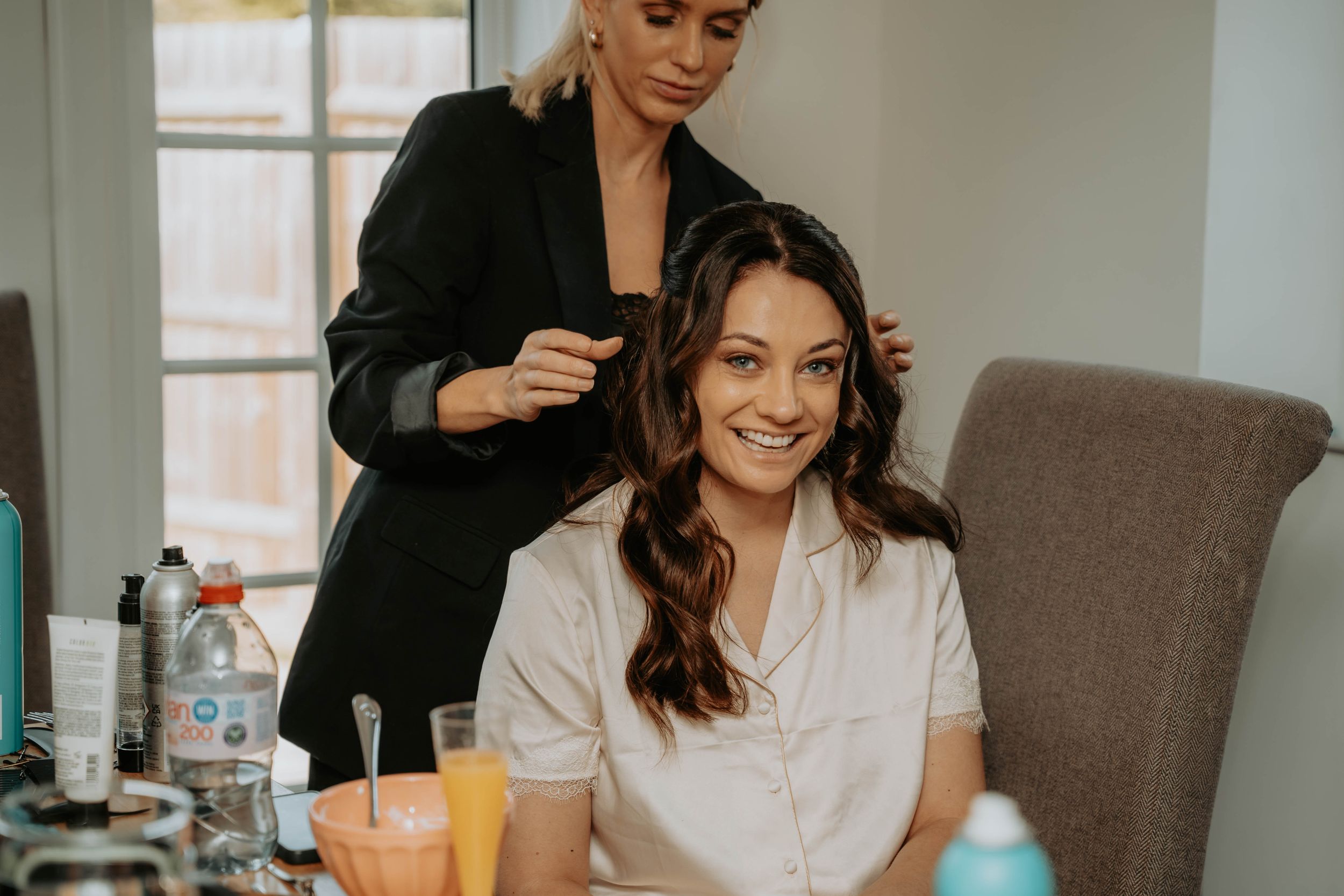 A hairstylist in a black blazer styling a smiling brunette woman's hair at a vanity table.