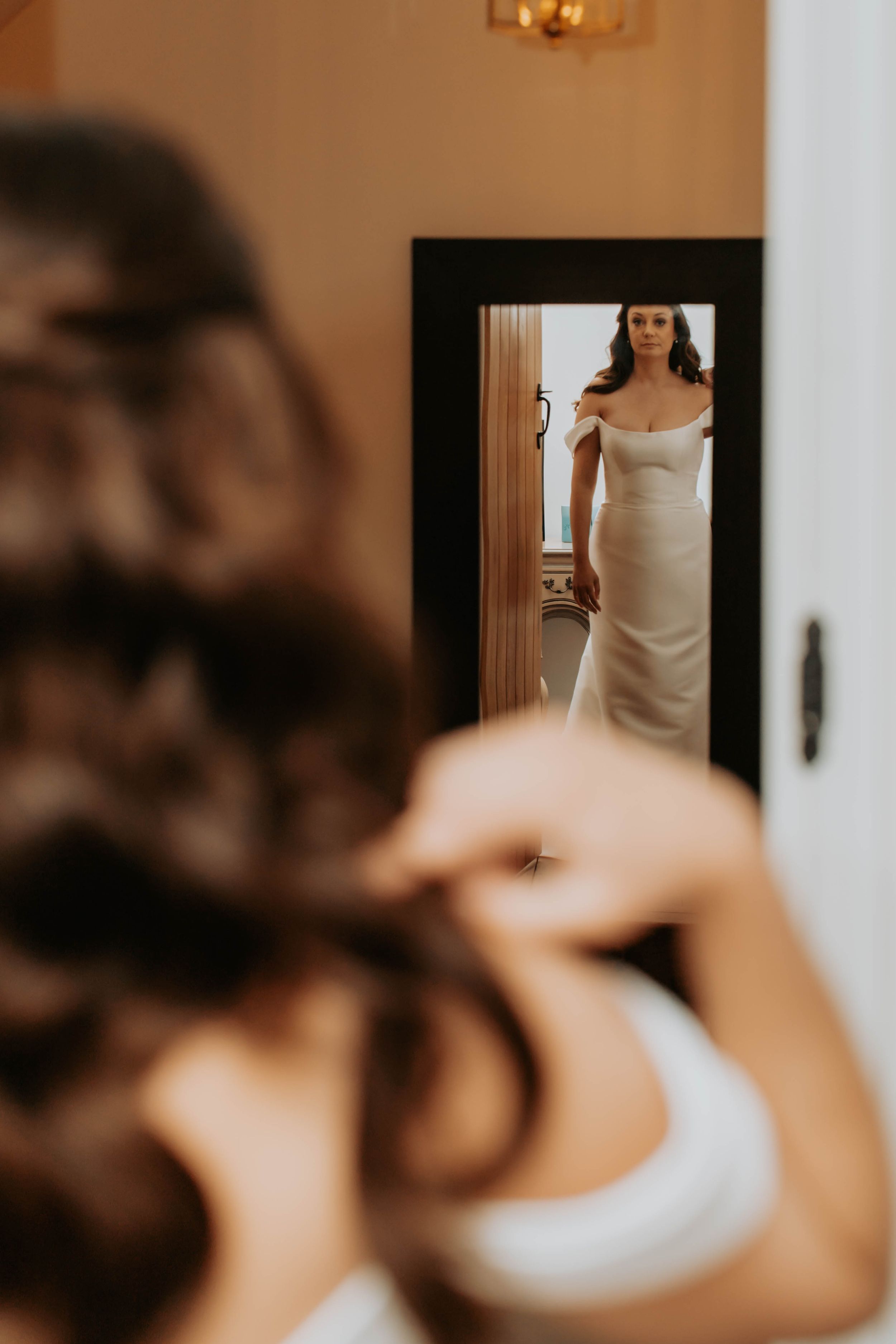 Bride in elegant white off-shoulder gown admires her reflection in a full-length mirror during wedding preparations.
