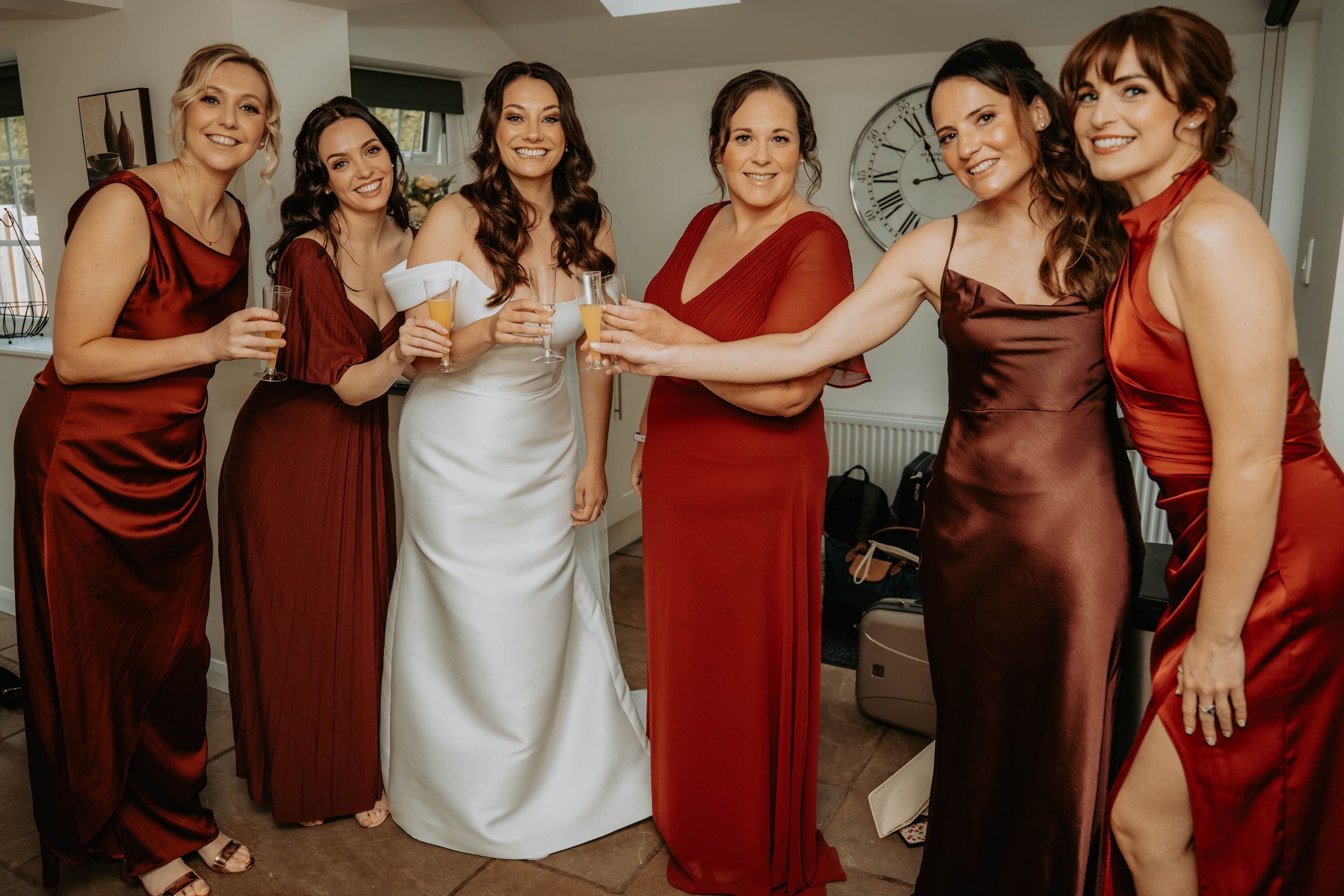 Bride in white gown poses with five bridesmaids in red and brown satin dresses, toasting with champagne glasses.