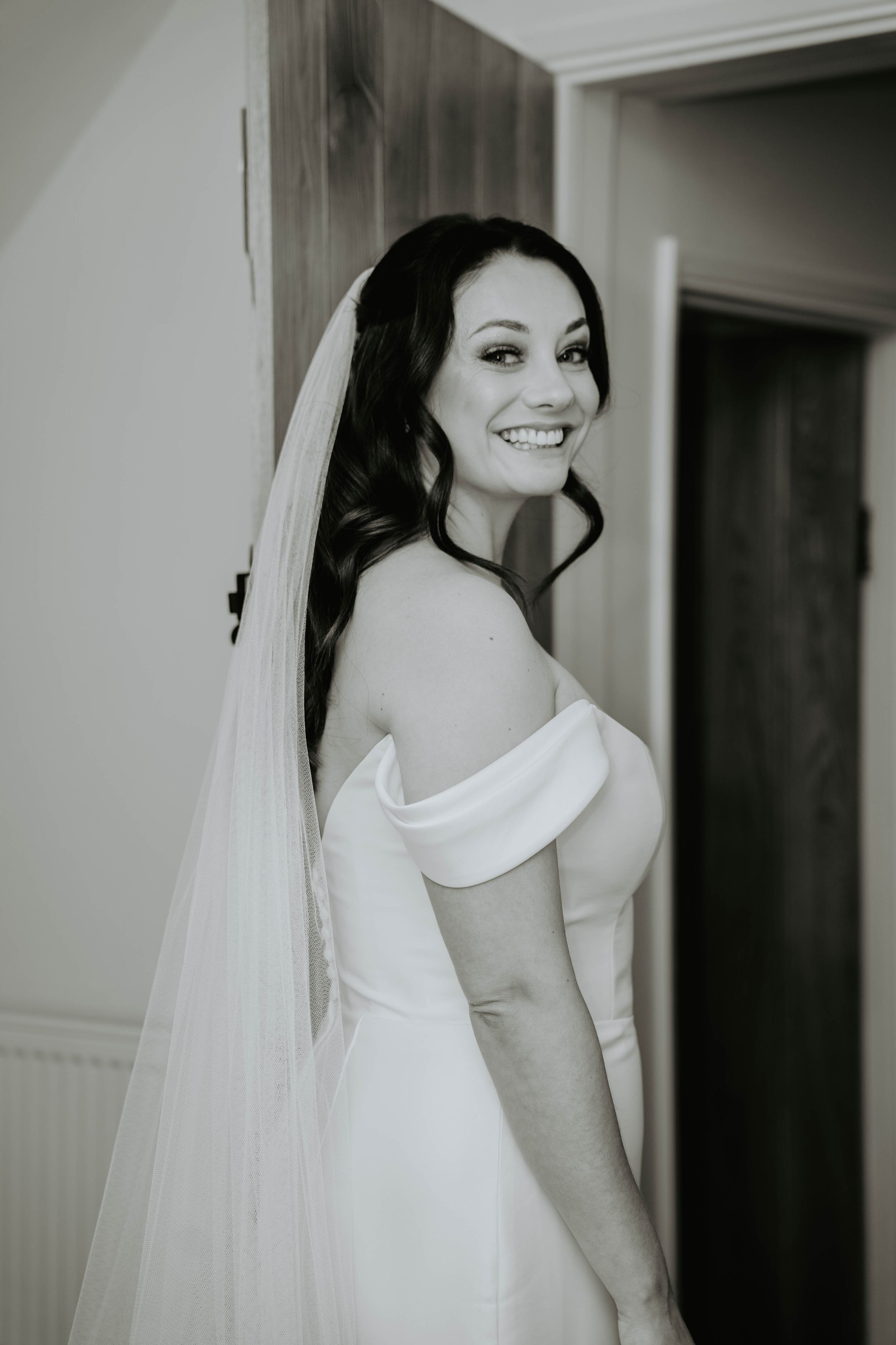 Smiling bride in off-shoulder wedding gown and veil poses elegantly in black and white portrait photo.