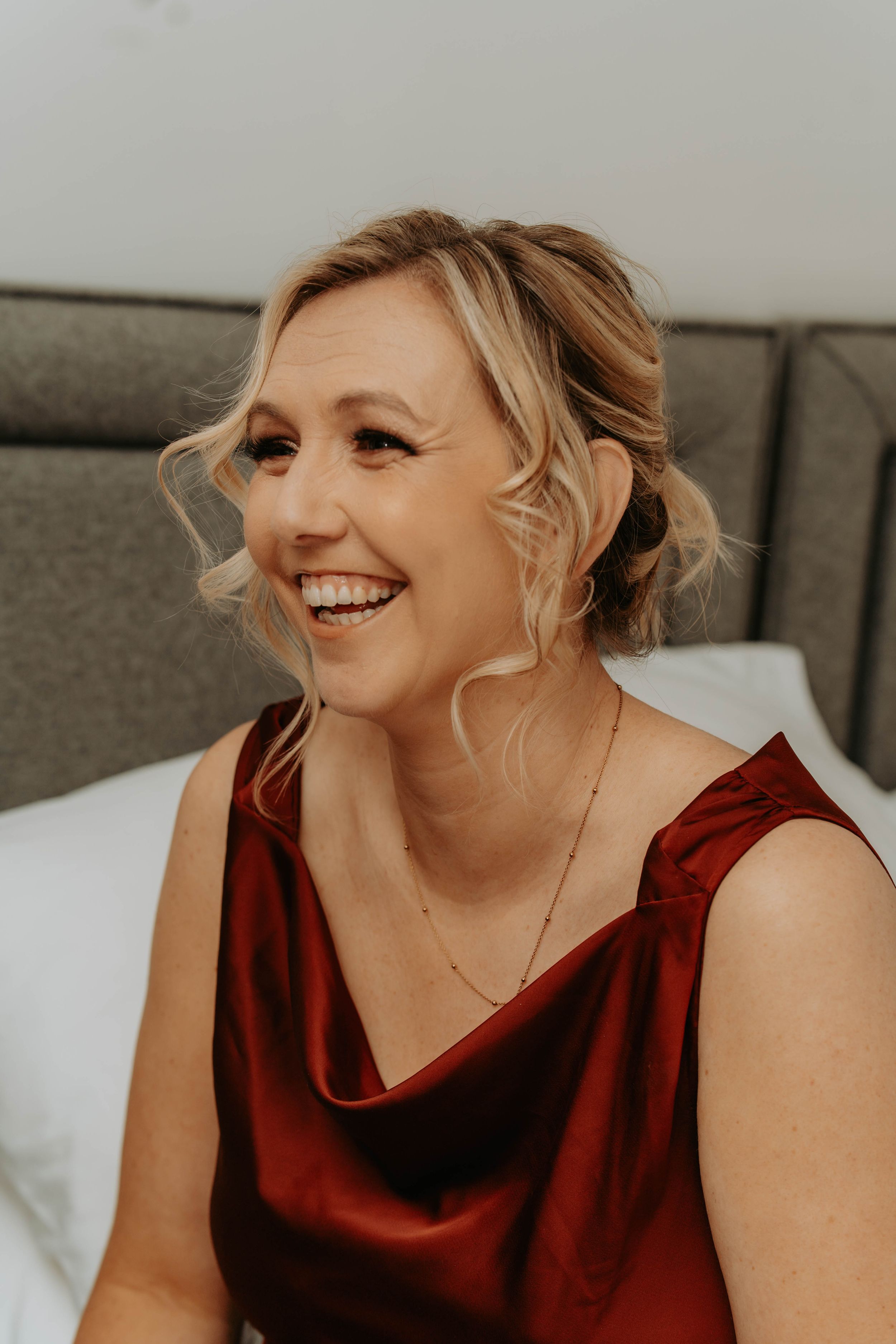 Smiling blonde woman in a red satin dress with an updo hairstyle, laughing joyfully indoors.