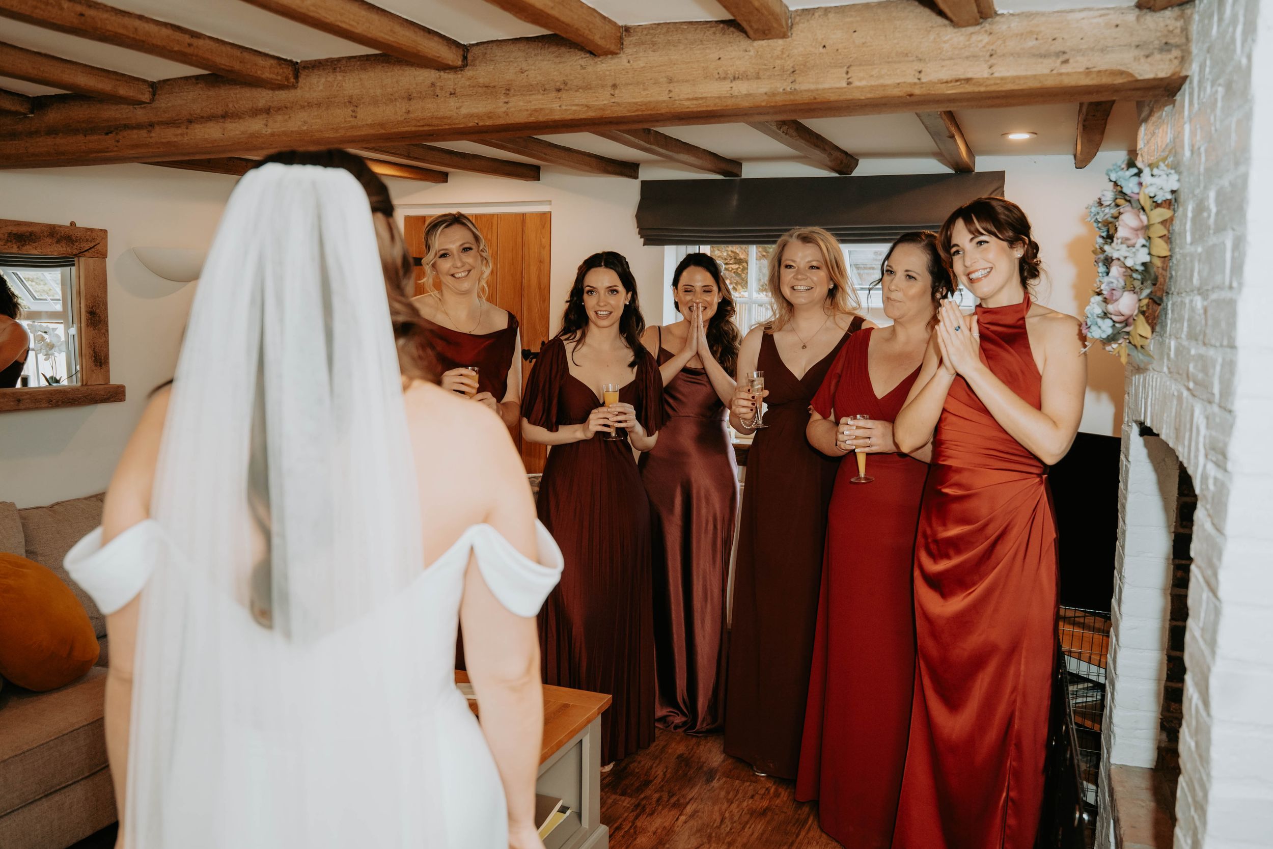 Bride in white gown and veil faces bridesmaids in red dresses holding champagne glasses during wedding preparations.