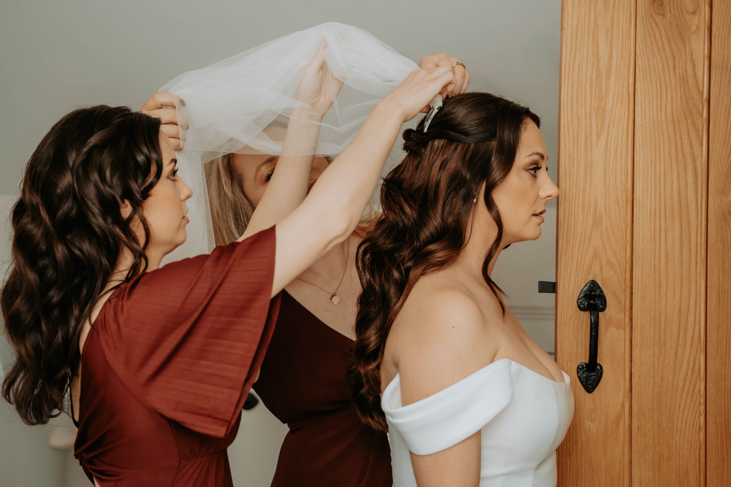 Two bridesmaids in burgundy dresses help a bride in a white off-shoulder gown with her veil before the wedding ceremony.