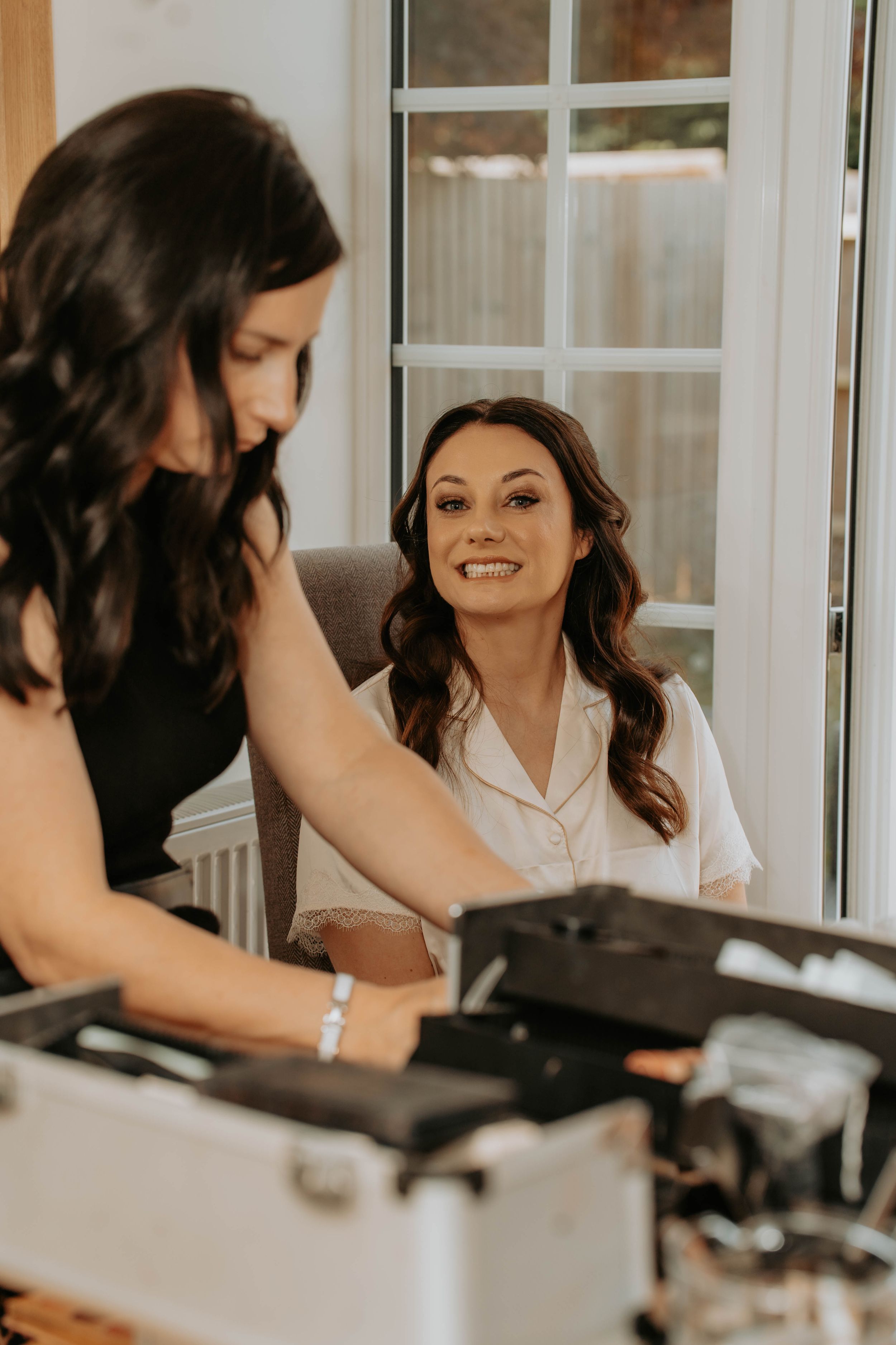 Bride smiling while makeup artist prepares cosmetics from a professional kit during bridal getting ready session.
