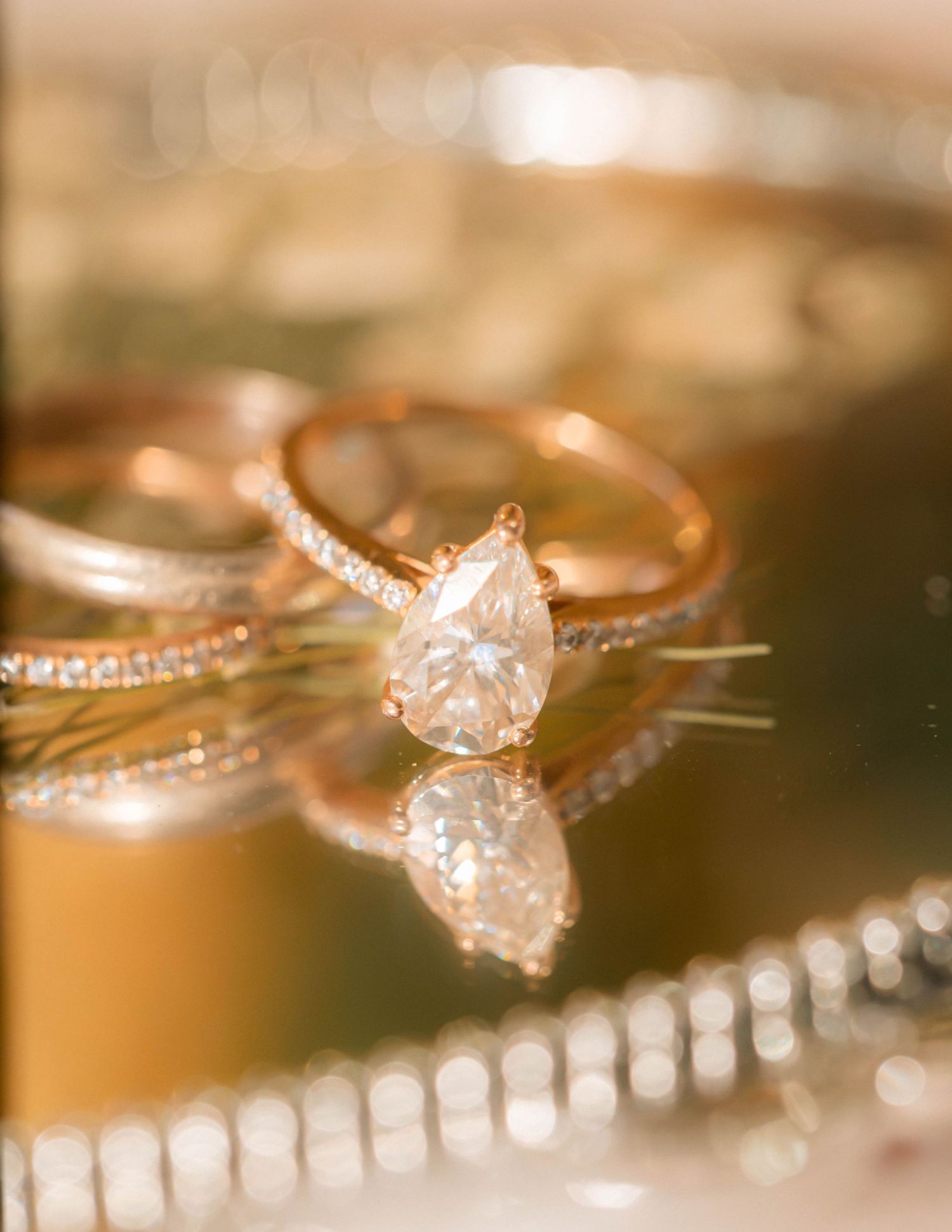 Rose gold pear-shaped diamond engagement ring with pavé band reflecting on a mirrored surface.
