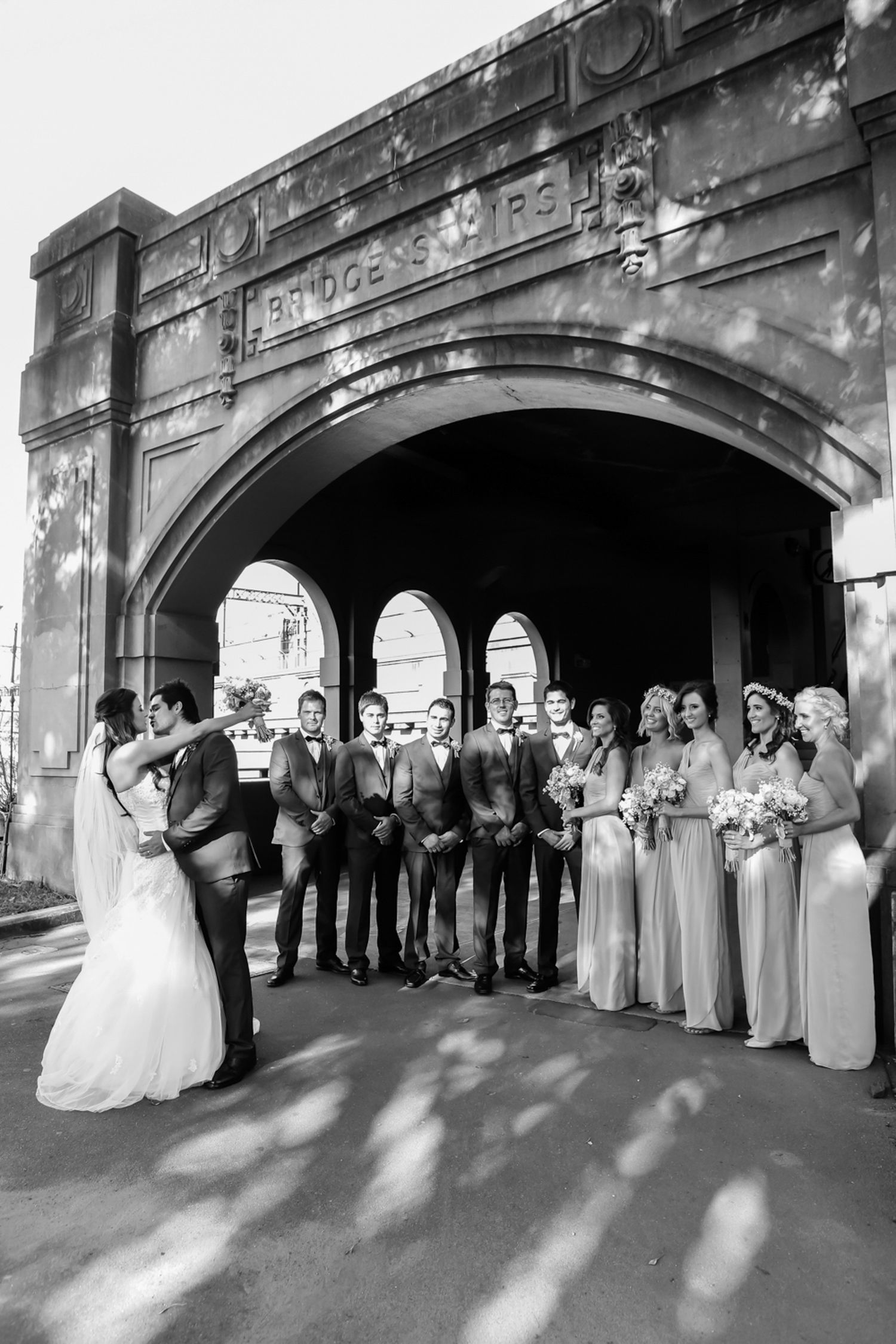 Black and white photo of bride and groom kissing under stone arches with wedding party, Bury St Edmunds wedding photographer.