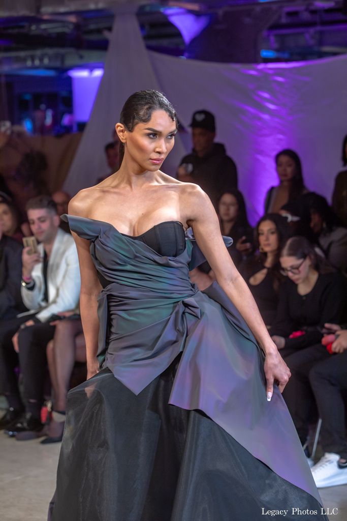 Fashion model in a sculptural gray dress photographed at a lively runway event.