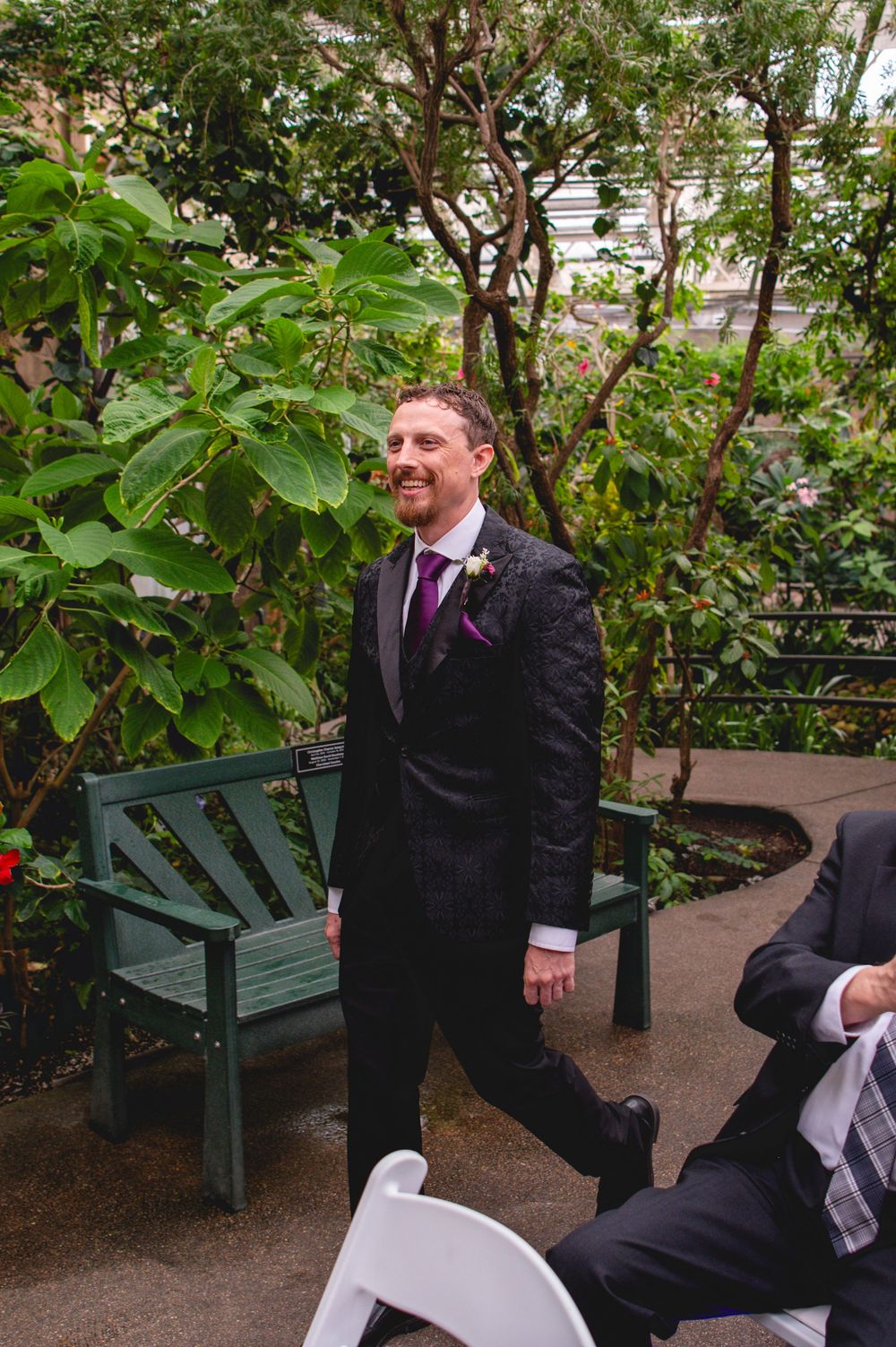 Smiling groom in dark suit with purple tie and boutonniere walks at outdoor garden wedding ceremony.