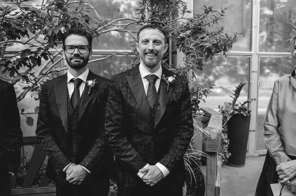 Two grooms smiling in black tuxedos at wedding ceremony in black and white photo.