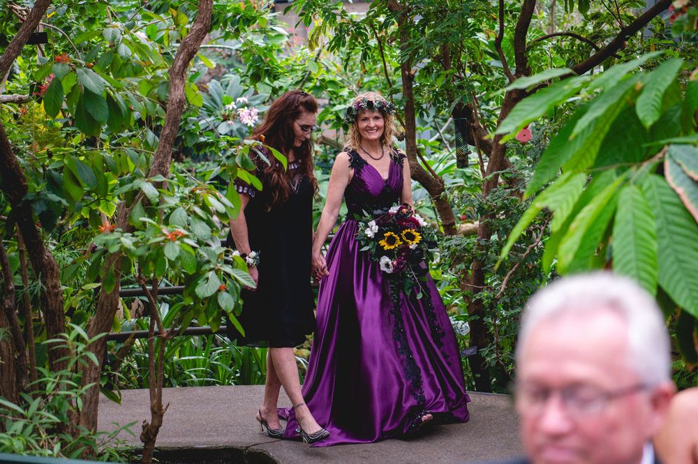 Bride in purple wedding gown with floral crown walks through lush garden path accompanied by woman in black dress.