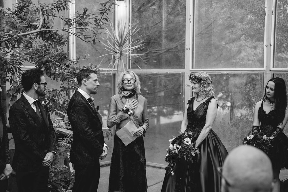 Black and white wedding ceremony photo with officiant, couple, and bridesmaids in elegant dark attire indoors.
