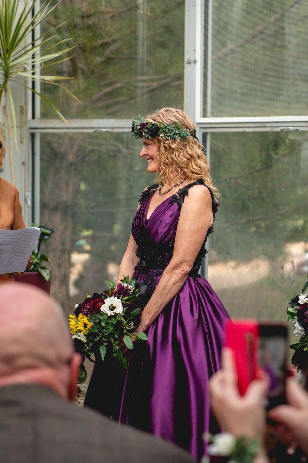 Woman in purple gown and floral crown smiles while holding bouquet at wedding ceremony.