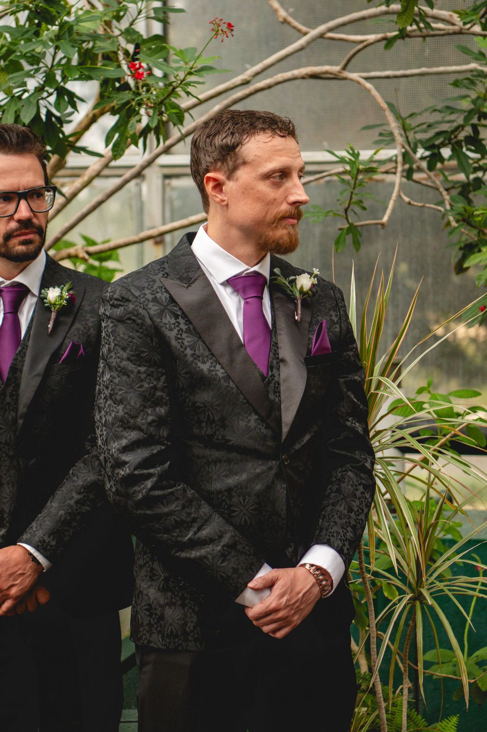Groom in black floral tuxedo with purple tie and boutonniere stands at wedding ceremony surrounded by greenery.