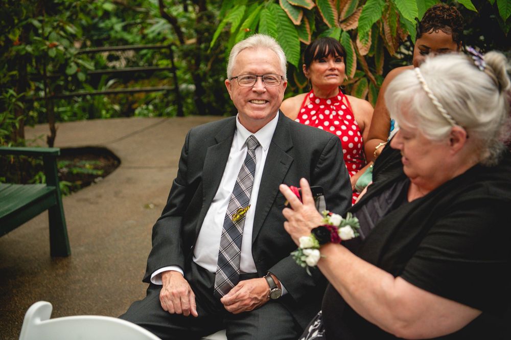 Elderly man in suit and tie smiling at outdoor event with guests in tropical garden setting.