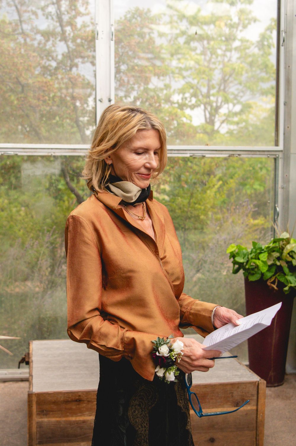 Smiling blonde woman in orange silk blouse holding papers, wearing floral wrist corsage in a greenhouse setting.
