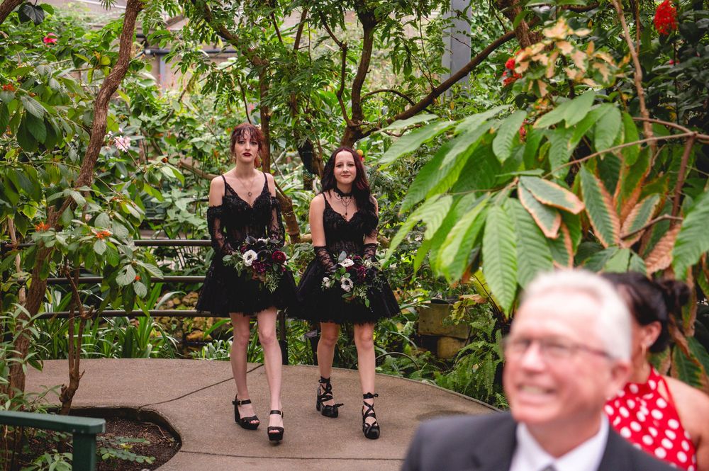 Two bridesmaids in black dresses carrying floral bouquets walk through a lush green botanical garden venue.
