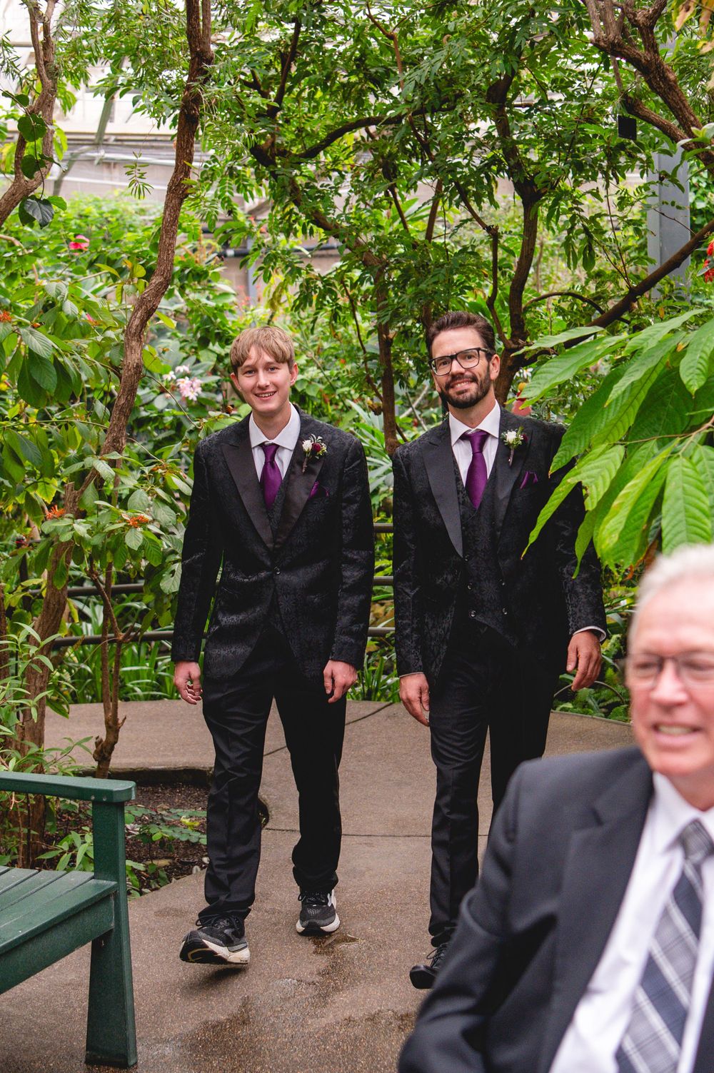 Two grooms in black suits with purple ties walk through a lush garden setting on their wedding day.