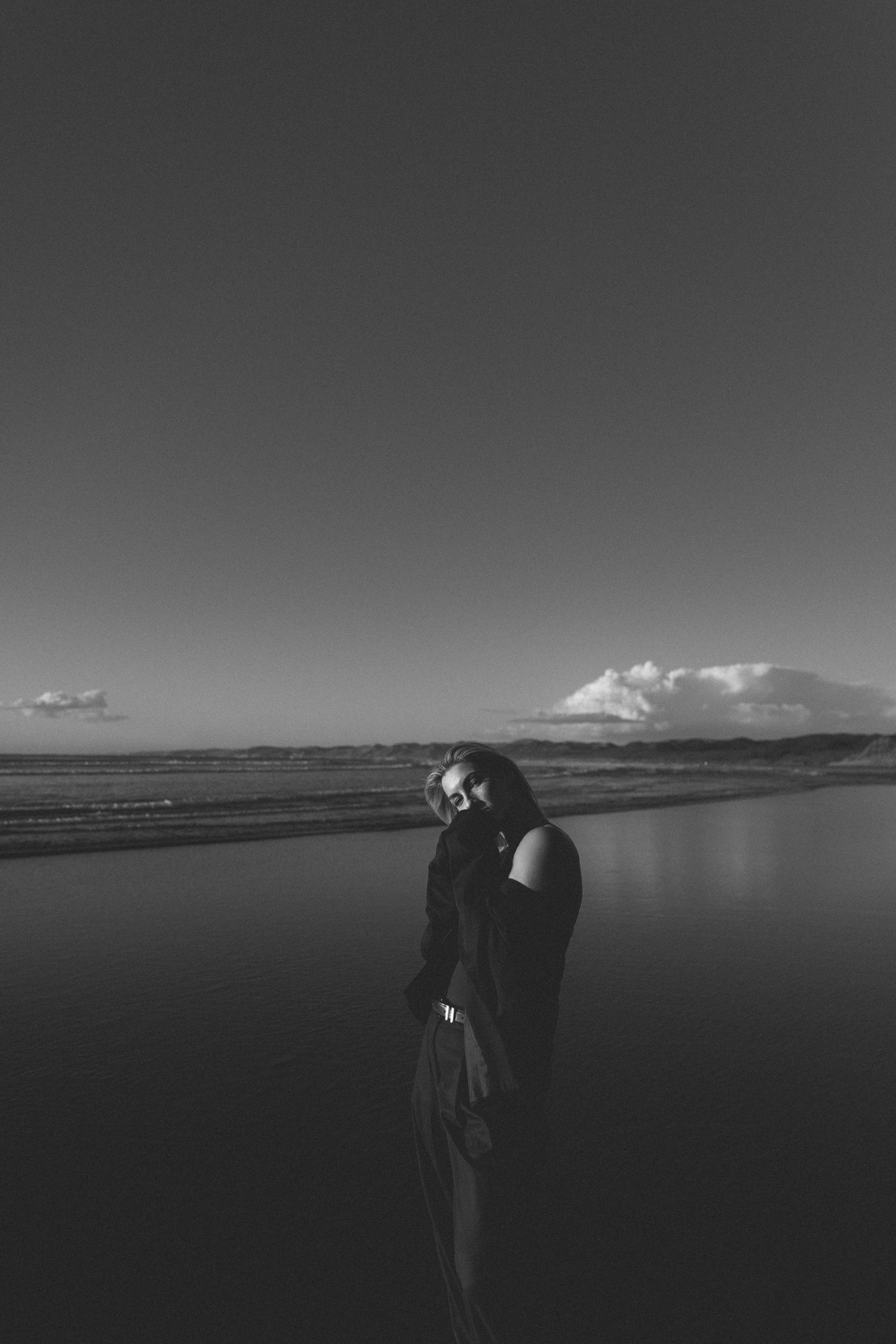 Black and white photo of a woman standing on a beach with ocean waves and clouds in the background in Raglan New Zealand