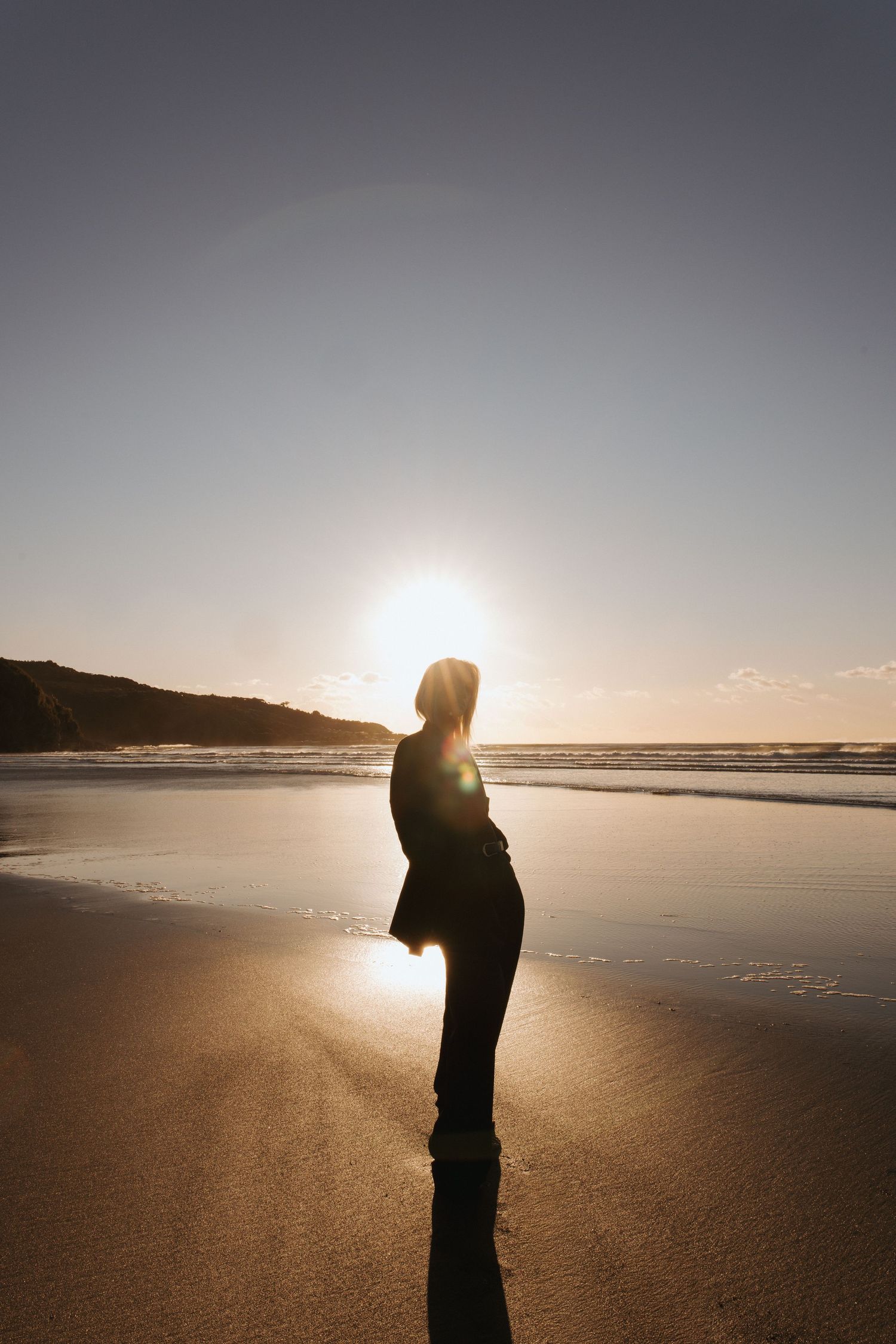Silhouette of a woman standing on a sunny beach at sunset with golden light reflecting on wet sand in New Zealand
