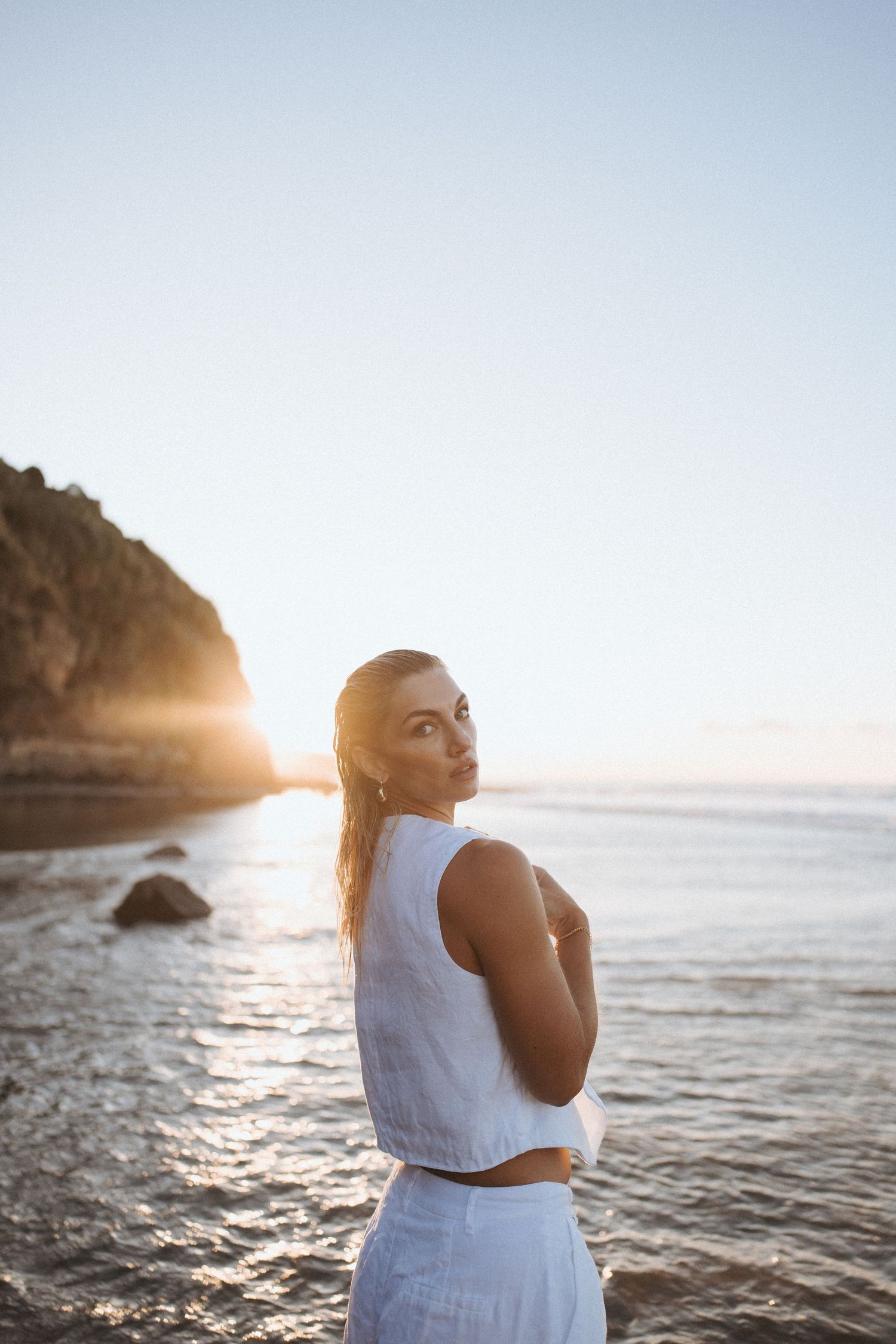 Woman in white outfit standing on a beach at golden sunset with rocky cliffs and shimmering ocean in background.