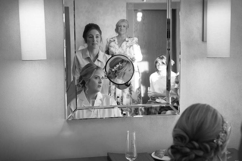 Black and white photo of bride getting ready with bridesmaids reflected in vanity mirror at Hetland Hall wedding venue