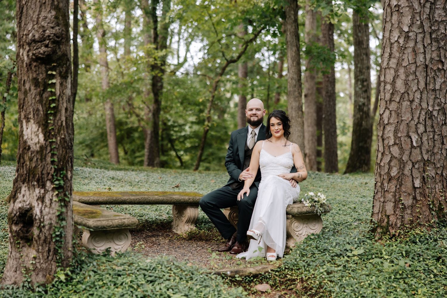 A bride and groom pose together on a stone bench surrounded by lush greenery in a wooded forest setting.
