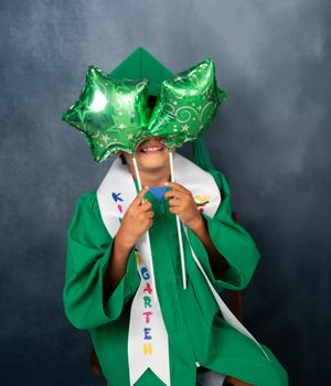 Child in green kindergarten graduation cap and gown holds star balloons covering their face, smiling for photo.