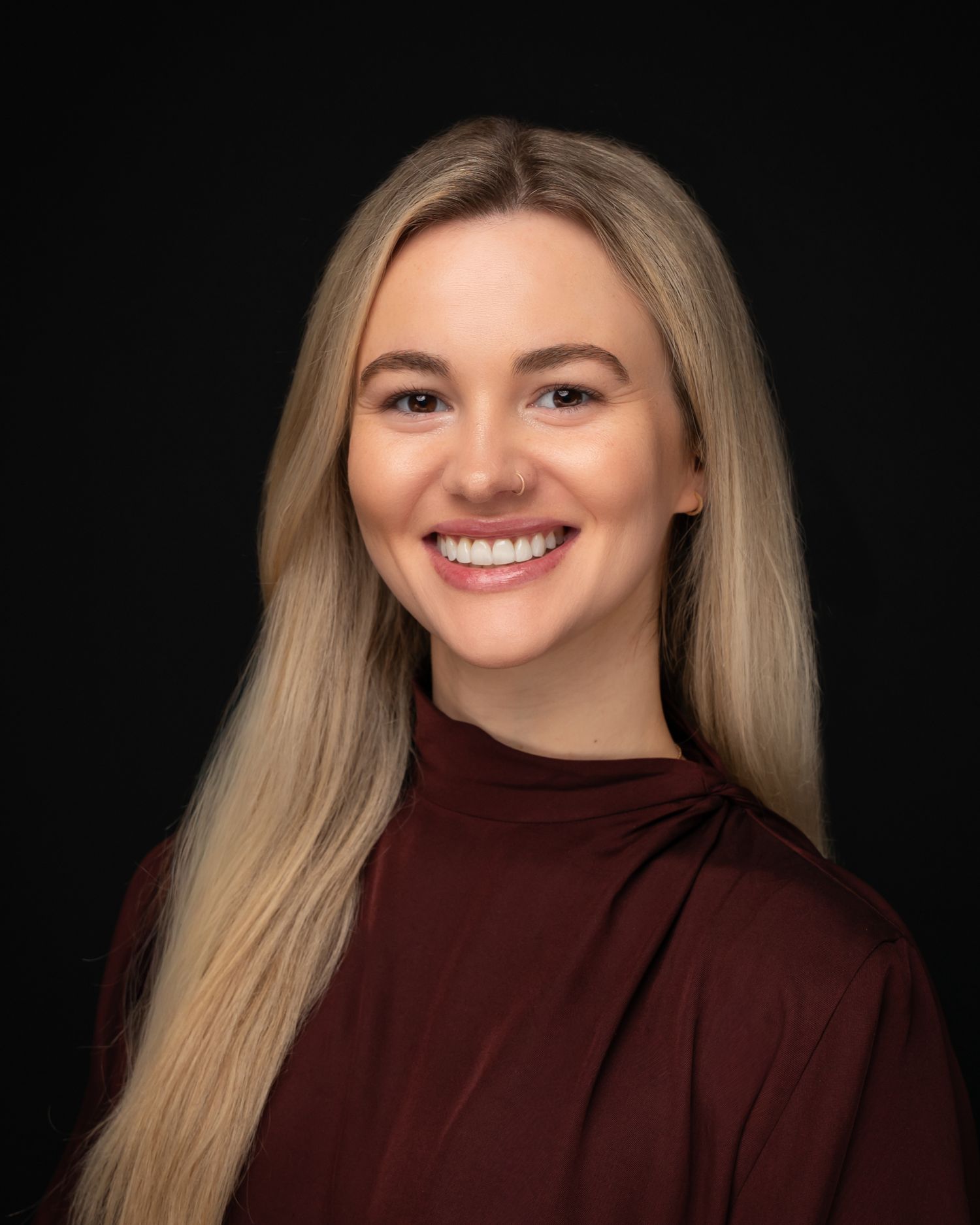 Smiling blonde woman in a dark red turtleneck top poses for a professional headshot against a black background.