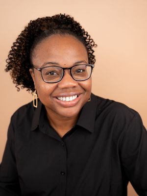 Smiling Black woman with curly hair, glasses, and gold earrings wearing a black shirt against a beige background.