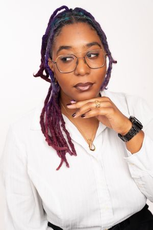 Woman with colorful purple and red locs wearing glasses and white shirt, posing thoughtfully against white background.