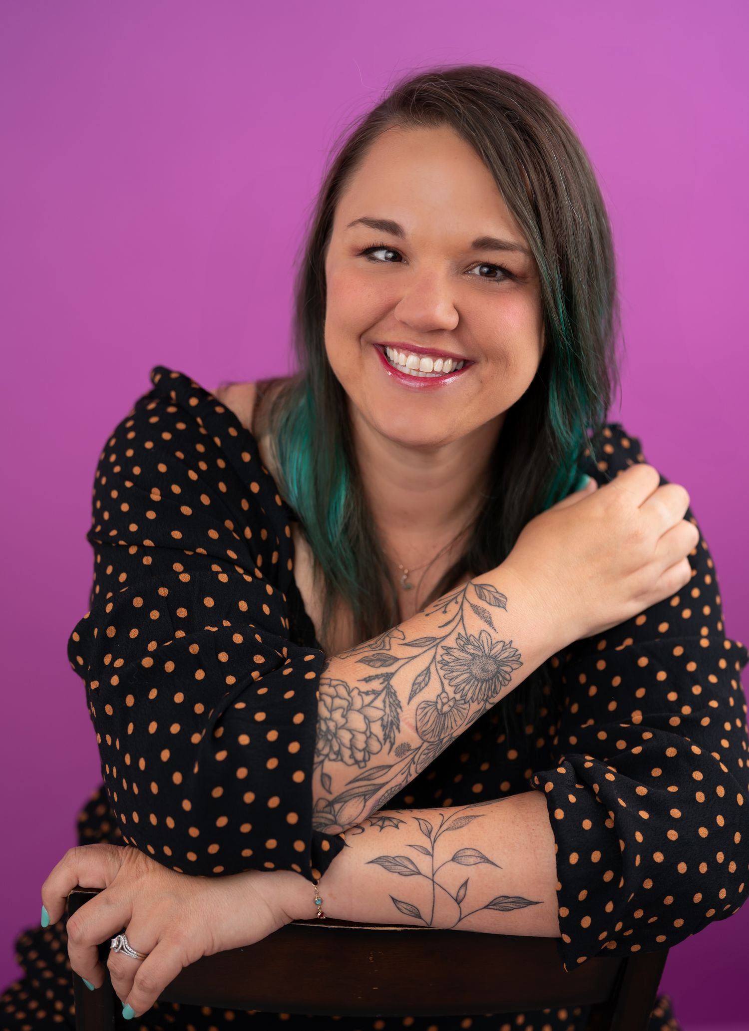 Smiling woman with teal hair and floral arm tattoos wearing a black polka dot top against a purple background.